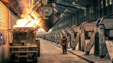 A view inside an area of the smelter called the potroom. An operator dressed in protective clothing uses a remote control to tip a large pot of molten metal into another pot, which is on the back of a trailer pulled by a tractor. 