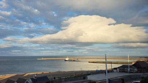 A sea view featuring a curved pier in the distance with a short jetty in front of it. A vessel, possibly a dredger, lies between them. In the foreground is a road and low buildings in front of a strip of sandy shore. The weather is sunny, with broken high cloud and a large flat cloud prominent in the foreground.