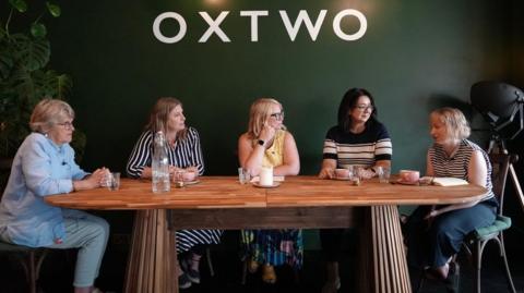 Five women are sat around a dining table.  There pink coffee cups, a candle, water tumblers and a bottle of water on the table.