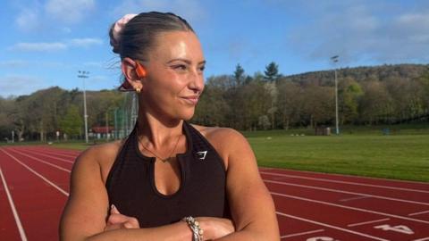 Alysia Mcandrew stands on a running track wearing a black running vest with her blonde hair scraped back in a bun and tied with a pink scrunchie. She is looking off to the side with a smile on her face.
