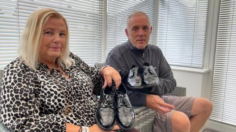 A woman with bobbed blonde hair, and a man with short grey hair, sit on a sofa holding two very similar pairs of patent leather girls' schools shoes.