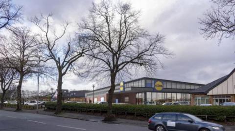 A computer generated image of the Lidl store next to retained units at Fallowfield Retail Park, with cars parked in front of the store, fronted by trees and bushes and a car is shown driving along the road in front of the site