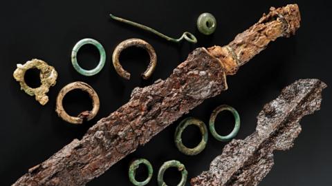 A selection of objects including bronze cloak pin, a glass bead pendant and toe rings on a black background