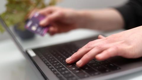A hand is typing on a laptop keyboard. The person typing is holding a purple bank card in their other hand.