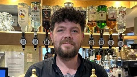 Jay Kemsley is standing behind the counter at a bar. He has short brown, curly hair and and a beard. Various spirits are hung up on the wall behind him.