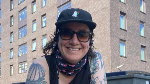 Hannah Cox wears a black baseball cap, sunglasses and neck scarf with a strapped running top. She smiles as she poses with a multi-storey buildign behind.