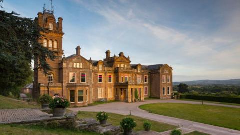 The front aspect of Hestercombe House, a grand, beige-gold stoned building that has a tower to the left. In front of the house is a large driveway with an oval grassy lawn. Fields and trees extend into the distance.
