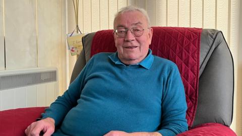 Murchadh MacIlleathain, air a bheil aillse a' phròstata agus e na shuidhe ann an cathair agus gàire air aodann. Tha geansaidh ghorm air agus plaide dhearg air a' chathair.