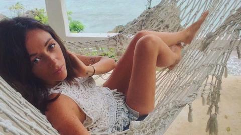 A young woman with long dark hair takes a selfie while sitting in a hammock. The ocean can be seen in the background. 