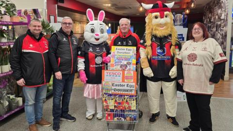 Three men, a woman, an Easter Bunny mascot and a Goole Vikings mascot standing in a line, looking at the camera. They are all dressed in red and black Goole Vikings uniform. They are stood in a supermarket. The man in the middle is stood behind a trolley full of Easter Eggs and clearly marked "East Egg Donation" with colourful signs.