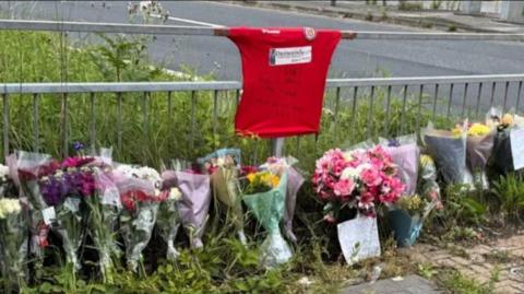 A wide shot of floral tributes laid for Israel Iyabor, which include a red t-shirt attached to crash barriers at the side of a road.  