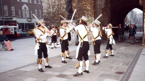 Five Morris men from King John's Morris Men wearing top hats white shirts, black trousers and white socks pulled up to the knee. They have yellow and black tassels and accessories. They are dancing in front of the bargate in Southampton at dawn 