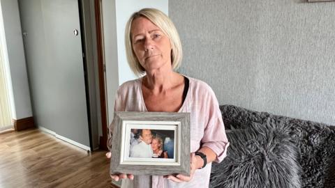 A woman in her fifties with blonde hair and a light pink top is holding a picture.  It shows her as a younger woman hugging her dad.  She is standing in her living room in front of a sofa.