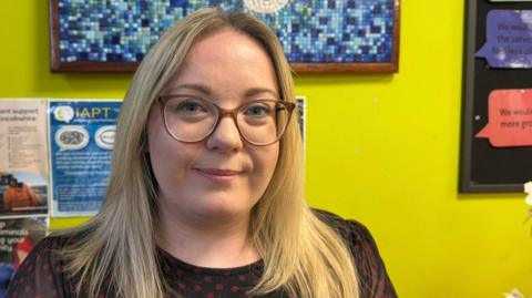 A woman smiling at the camera. She has blonde hair and glasses. She's standing in front of a wall painted yellow. Posters, slogans and a blue mosaic are attached to the wall. 