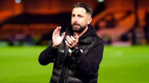 Kevin Horlock, manager of Maldon & Tiptree FC, stands on a football pitch, clapping. He is wearing a black hoodie with a black gilet over the top. He has short black hair and a black beard