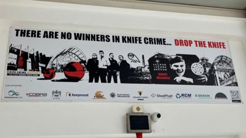 A black, white and red poster is on the wall above a door at Meadowhall. It shows images of Sheffield including Meadowhall, the Winter Garden and Kelham Island Museum