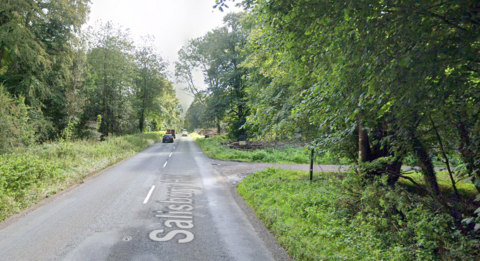 A main road through a forested area with a turning on the right and cars both approaching and driving away from the camera