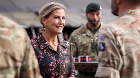 The Duchess of Edinburgh smiling at a soldier in a parade with her hair ted back and hands clasped together