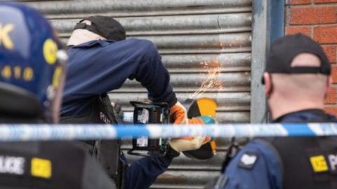 Police are carrying out a raid on the property on Lockett Street. Three police officers are facing towards a metal roller shutter - one is using angle grinder with sparks flying into the air.
