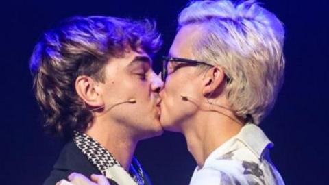 A well-lit couple kiss on stage against a dark background. Linus - who is wearing a flamboyant white shirt with an oversized half collar on the right hand side and dark rimmed glasses - has blonde hair combed in a quiff. Joseph is wearing a short navy jacket which has a large collar and a white, grey and black neck scarf. He has brown/ blonde hair.