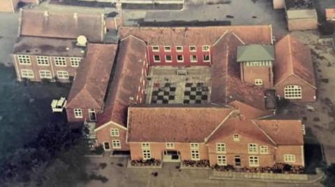 Aerial view of The Red House at Buxton, Norfolk, featuring a quadrangle of red brick buildings and ancillary buildings. There is a chequerboard pattern in the centre of the quadrangle, and greenery to the lower left of the image, and hard standing on all other elevations.