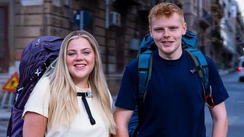 Katie wears a white t-shirt and blue backpack. She has long blonde hair and Harrison has short ginger hair and wears a blue t-shirt and blue backpack with a red hook on the front 