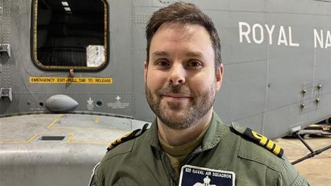 Dressed in a green shirt with a name label Commander Alasdair "Al" Woodward CO of 820 Naval Air Squadron stands in front of a Merlin, like the aircraft which flew to Cyprus.