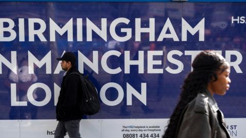 A blue sign with white writing reads "Birmingham Manchester London" as a man and a woman walk past in opposite directions in front of it