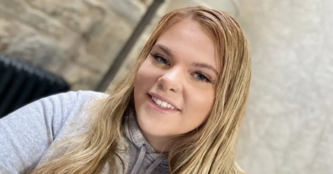 Selfie of Bethany Branson, a teenager with long blonde hair, blue eyes and long eyelashes. She is wearing a grey hoodie and the background, a wall, is blurred.