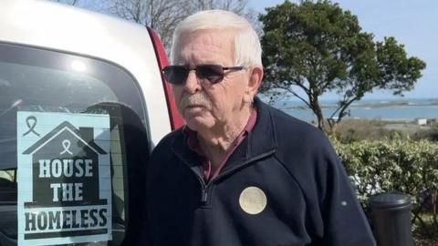 Man with white hair wearing sunglasses in a navy quarter-zip jumper standing next to a van with a sign that says 'house the homeless'.