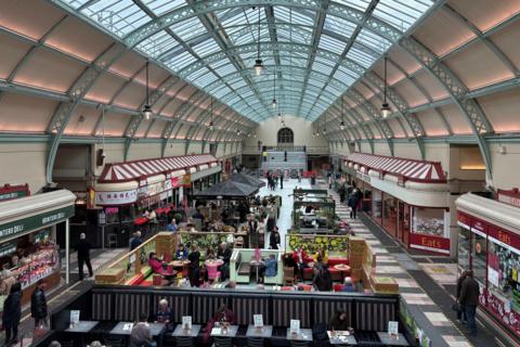 Inside Grainger Market. Seats for diners and shoppers are placed between rows of freshly painted stalls. The glass barrel roof runs the length of the room. There is a large open space leading to a staircase at the far end.