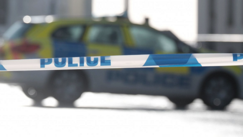 A police cordon tape with blue writing with a police car out of focus in the background.