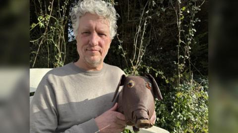 A man with white, curly hair in a grey jumper. He is holding a leather doll which looks like a dog with a pig's nose.