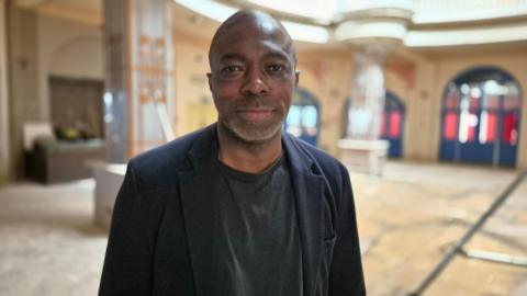 A picture of Bayo Alaba, standing indoors and wearing a black jacket and black t-shirt