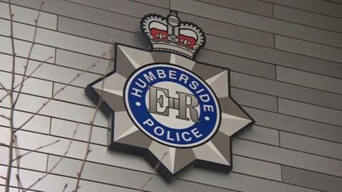 The Humberside Police logo - featuring a blue circle, a silver star and a red crown - on the side of a building. A tree branch is in the foreground of the image and the building is covered in grey cladding