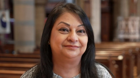 Sbba Siddique is stood in the church, with the pughs in the background, and is wearing a white and black top. She has long, black hair.