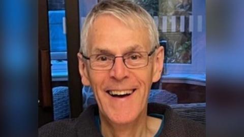 Andrew Phillips has grey hair and glasses and is smiling at the camera. He is wearing a dark jumper with a blue T-shirt underneath.