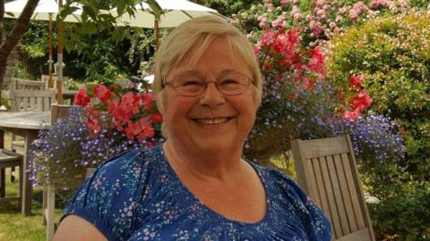 79-year-old Gwynneth Exall wearing a blue and floral top and is sat smiling, wearing glasses with grey short hair. 