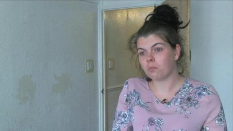 A woman with tied back hair and a pink top looks fraught with a bare white wall behind her. 