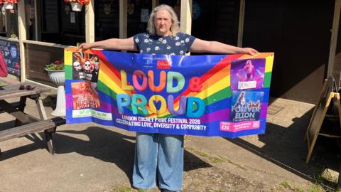 A woman stands outdoors holding a colourful “Loud & Proud” Pride festival banner in front of a wooden building with hanging baskets and string lights.