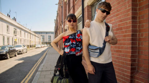 A woman and a man stand in a terraced street in sunglasses with a duffel bag, a red, navy and blue corset and a small man bag made of seat belts
