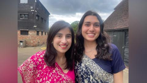 Two young women pose for a photo in an outdoor area - both are wearing colourful sari-like garments known as garas.