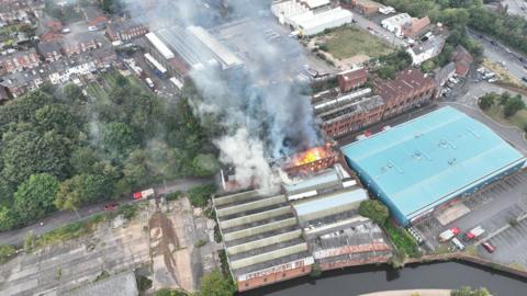 A drone photo of a warehouse building on fire, with canal in foreground, a large blue-roofed building on the right and homes and trees to the left. Fire and smoke is billowing from the warehouse roof.