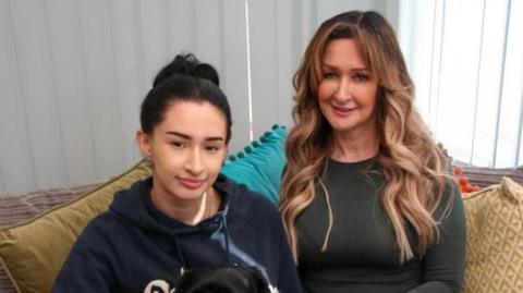 Two women are sitting on a beige-coloured sofa with yellow and blue cushions. The woman to the left is wearing a navy hooded jumper and has dark hair tied back. The woman to the right is wearing a grey long-sleeve T-shirt and has long blonde curled hair.