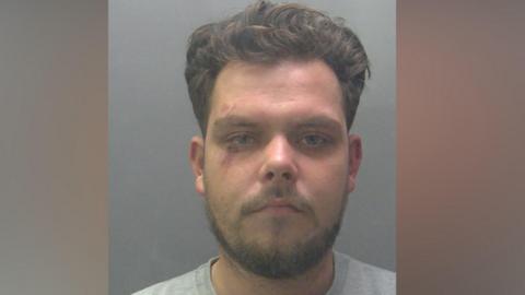 Custody image of Sonny Loveridge, who has brown hair and a short beard. He is wearing a grey sweater and has cuts surrounding his right eye.