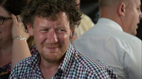 A man with curly brown hair, reddish facial hair, a small hoop earing in his left ear, and wearing a red and green checked shirt, smiles looking away from the camera.