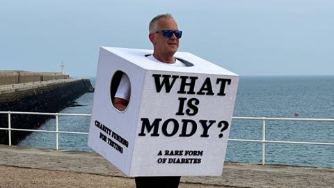 Richard is running long a seaside promenade wearing a large white cube costume with the text ‘WHAT IS MODY? A RARE FORM OF DIABETES,’ with the ocean and a pier behind him