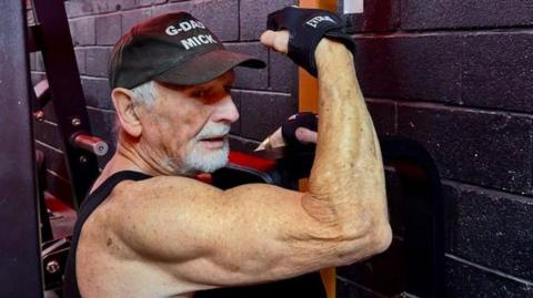 Mick Wadge weighlifting in a gym. His arm is lifted and he has toned biceps.