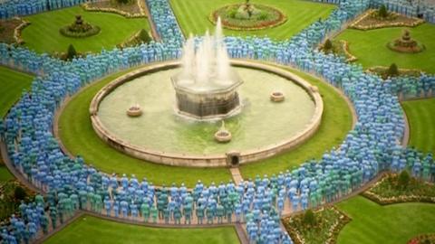 A sea of naked men and women stood in a circle around a circular water fountain. They are painted in several shades of blues and greens, and surrounding them is lush green grass and flower beds.