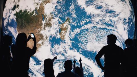 A lit sculpture of the Earth, with people silhouetted in front of it, pointing at it and taking photos.